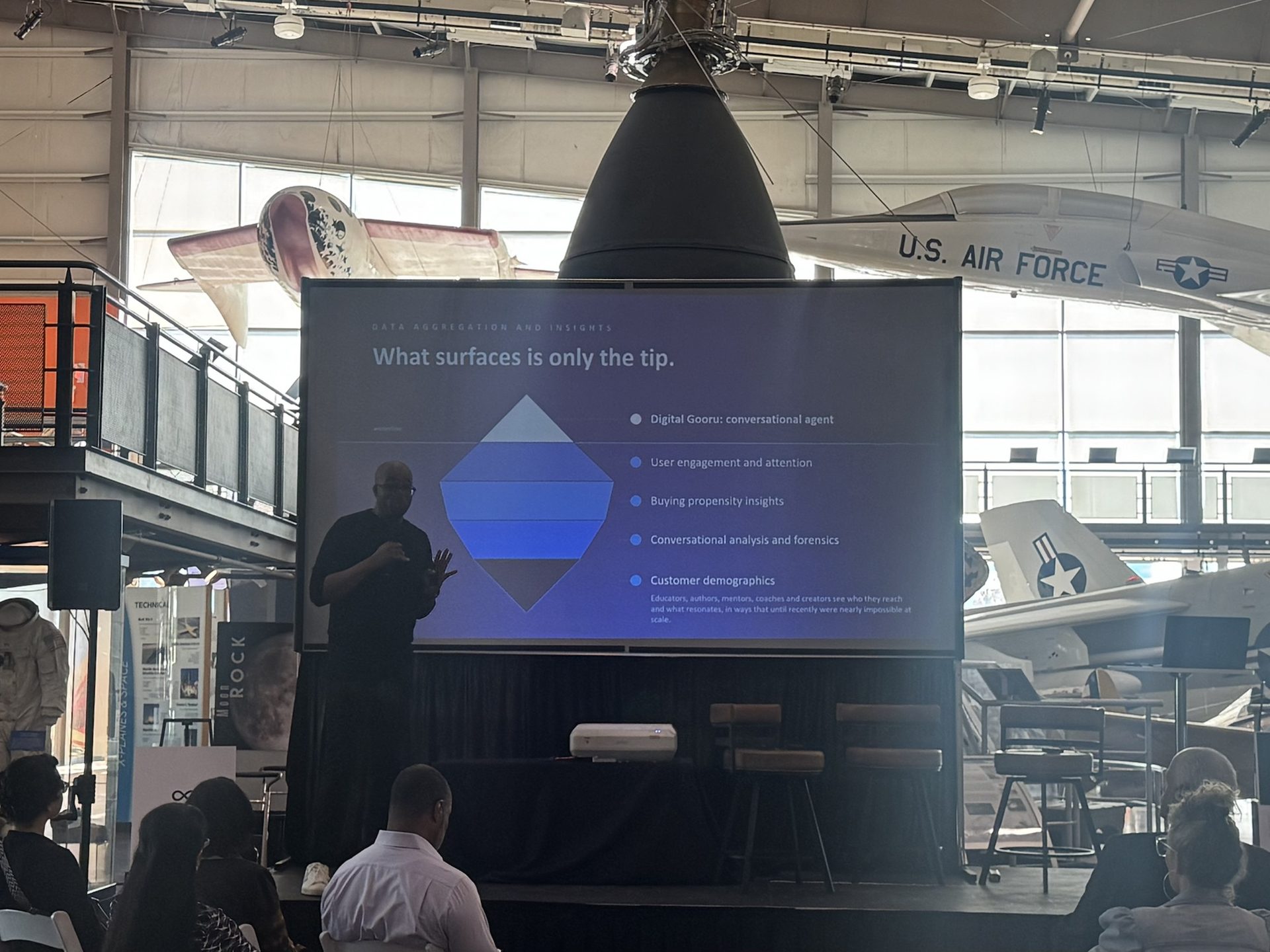 A presenter on the keynote stage with a projected slide holding its contrast against the daylit windows of the Frontiers of Flight Museum hangar