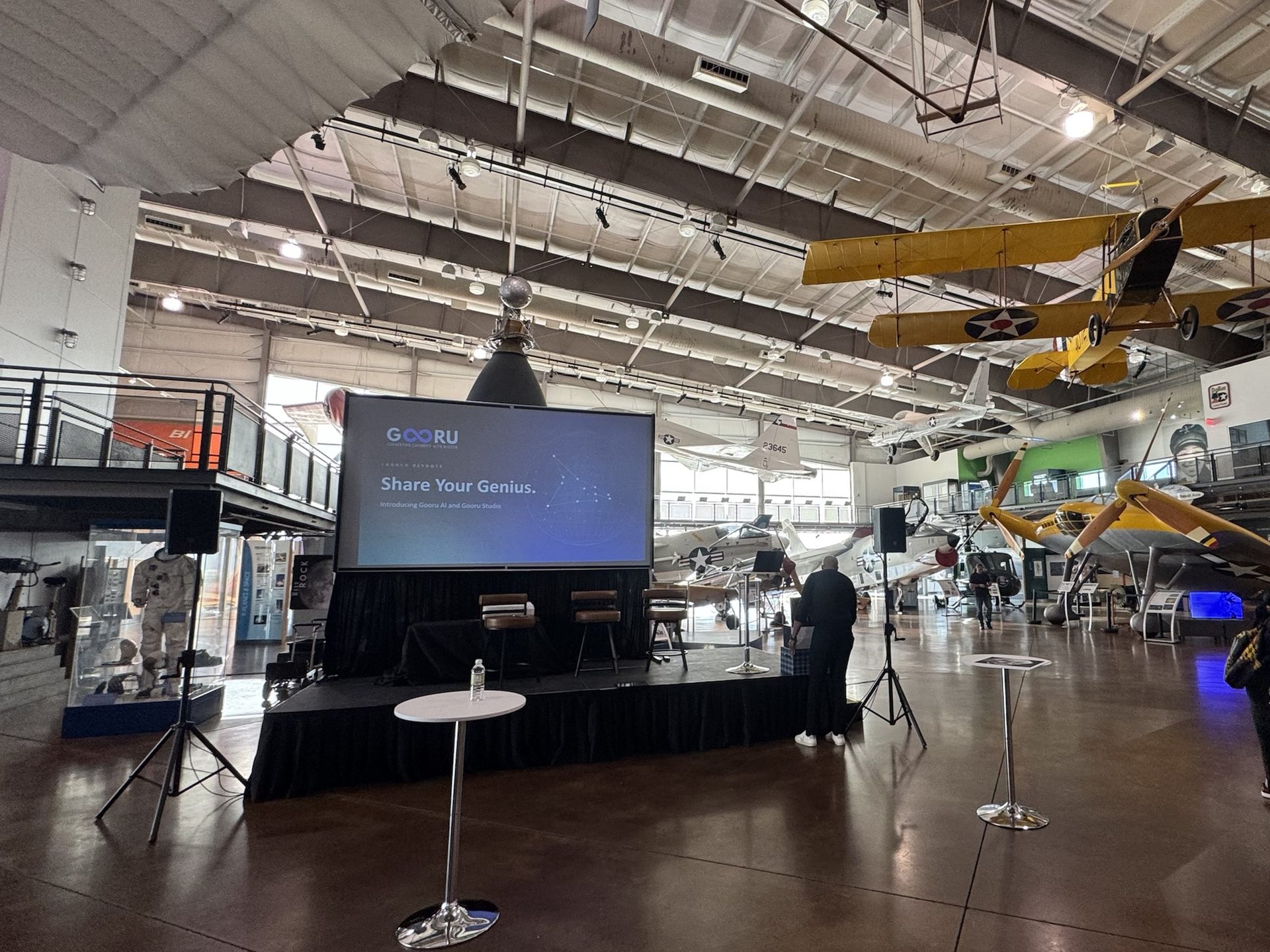 Keynote stage set before doors in the main hall of the Frontiers of Flight Museum, with a projection screen, an ultra short throw projector on the stage and speakers on tripods under hanging aircraft