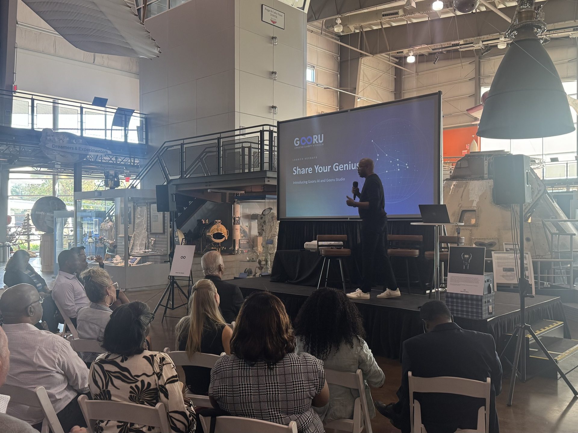 Audience seated in the Frontiers of Flight Museum hangar watching a product launch keynote, with a speaker on a tripod beside the stage and aircraft exhibits around the room