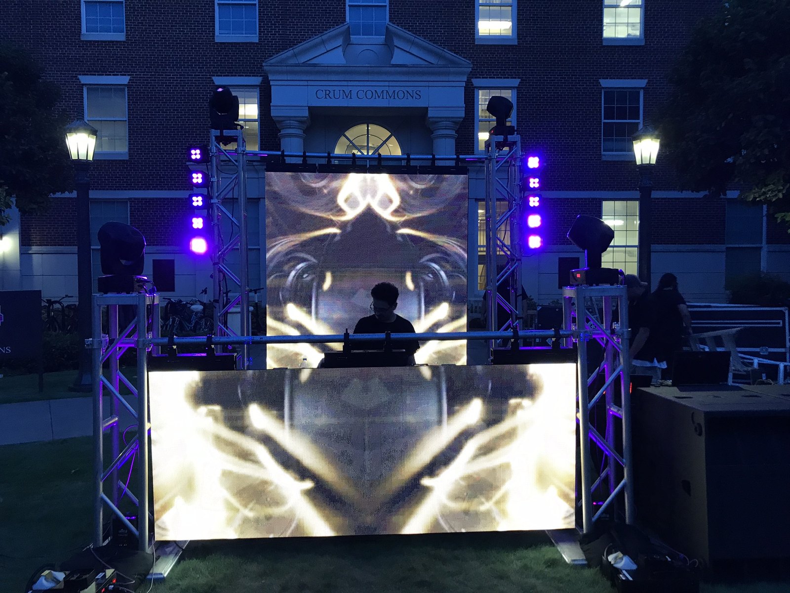A DJ booth lit from behind by an LED wall, framed by truss towers outside a brick campus building at night
