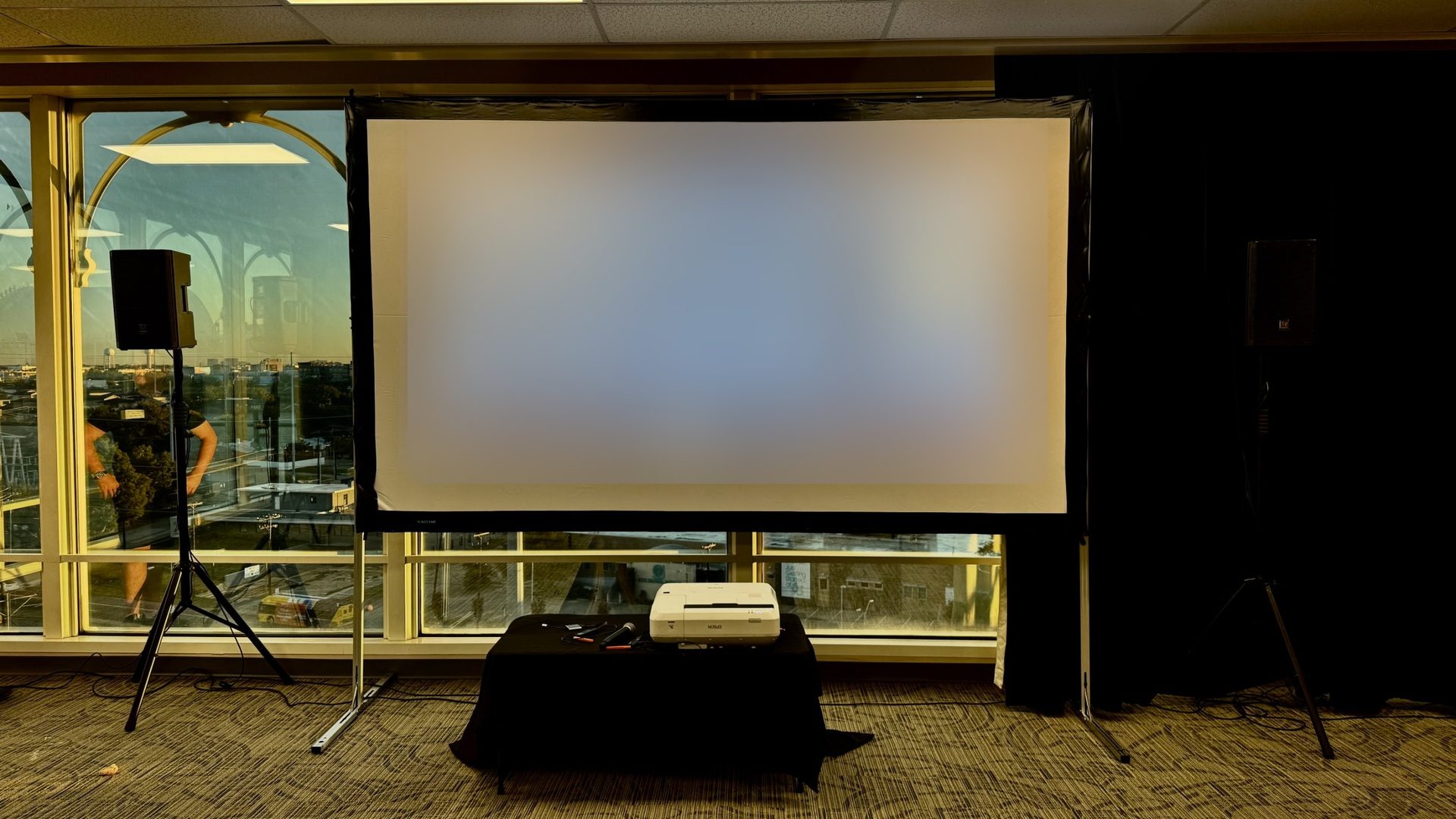 A corporate training room at the Infomart in Dallas with an Epson projector on a draped table, a framed projection screen and two EV ZLX speakers on stands in front of tall windows