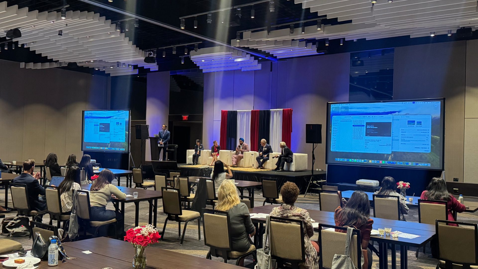 A medical symposium panel on a low stage in a Plano hotel ballroom, with two ambient light rejecting screens and BassBoss DV12 speakers on stands