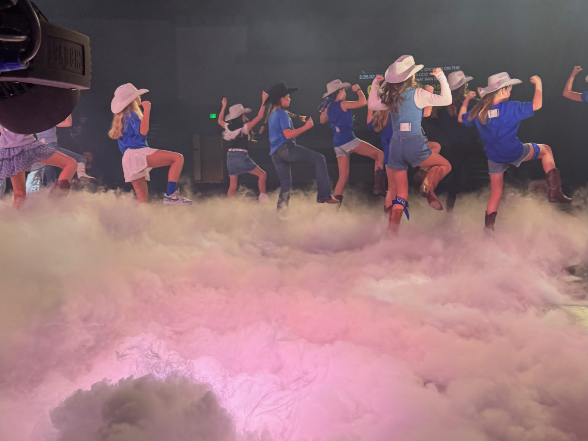 Children in cowboy hats dancing on a church stage above a thick layer of dry ice clouds lit pink