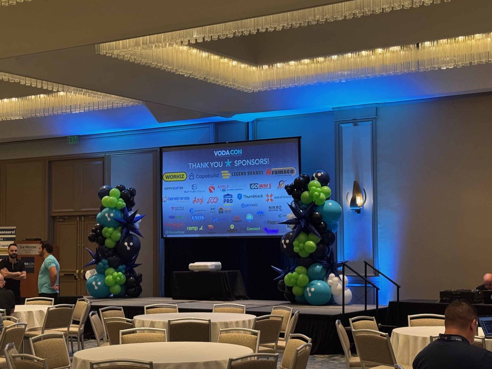 A conference stage in a Dallas hotel ballroom with a projection screen, blue uplighting and balloon columns