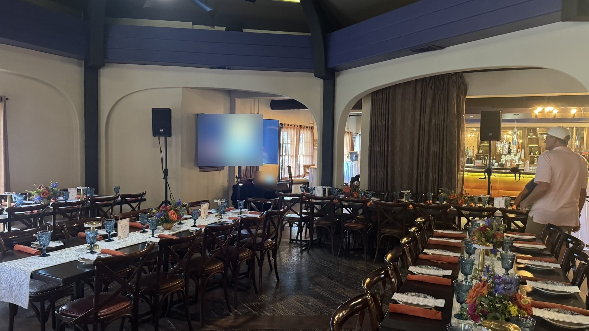 A 65 inch TV on a floor stand between two speakers on stands at the front of a private dining room set for dinner